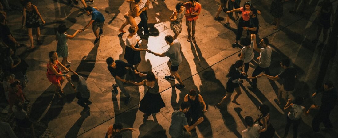 Seen from above, dozens of couples dance in pairs on a stone square at night, casting long shadows under the lights