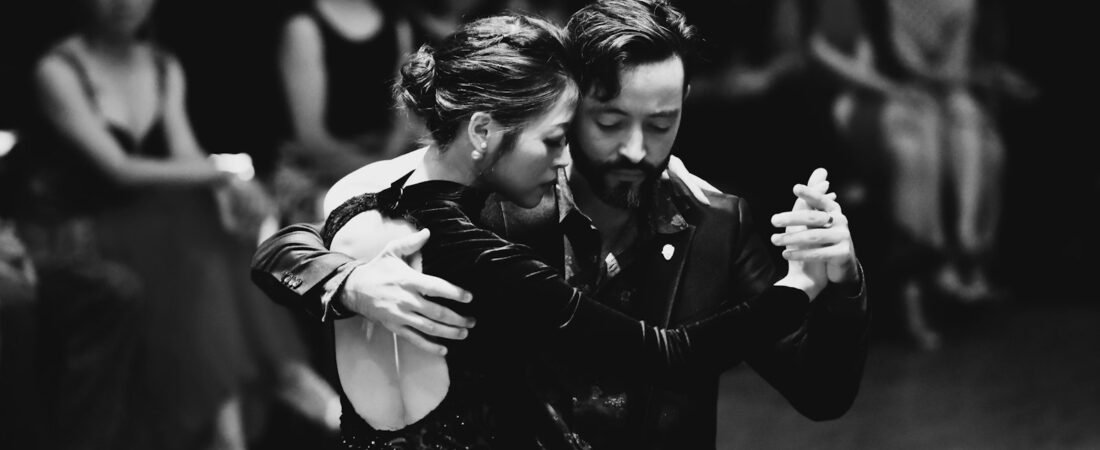 Two dancers in close embrace on a dance floor, with seated onlookers watching from the edge of the darkened room