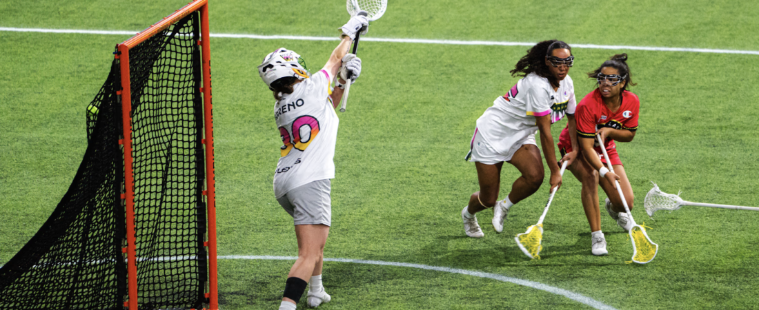 A lacrosse goalkeeper raises her stick beside the orange goal frame as two opposing players close in across the green turf