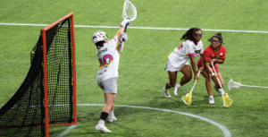 A lacrosse goalkeeper raises her stick beside the orange goal frame as two opposing players close in across the green turf