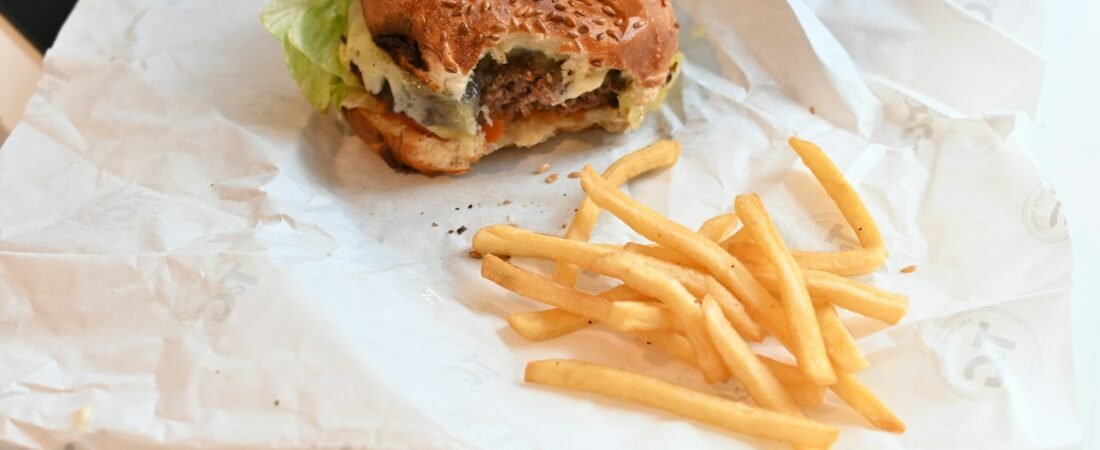 A single cheeseburger with a bite taken out of it, beside a small pile of french fries on white wrapping paper