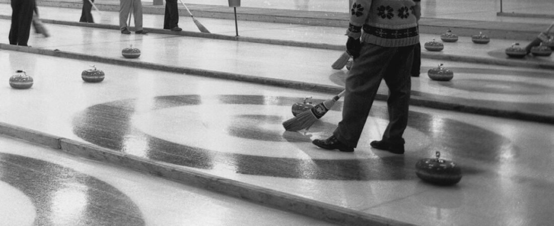 Curlers on several adjacent sheets of ice, a man sweeping with a corn broom in the foreground and about a dozen curling stones resting on the painted houses