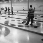 Curlers on several adjacent sheets of ice, a man sweeping with a corn broom in the foreground and about a dozen curling stones resting on the painted houses