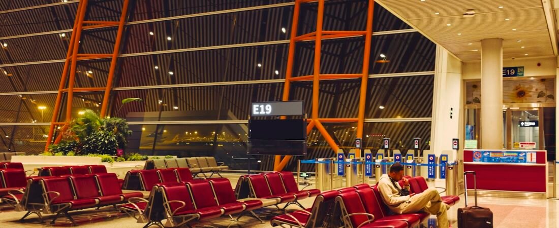 A lone traveller sits with a rolling suitcase among rows of empty red seats at an airport gate at night, below a blank gate display marked E19