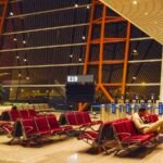 A lone traveller sits with a rolling suitcase among rows of empty red seats at an airport gate at night, below a blank gate display marked E19