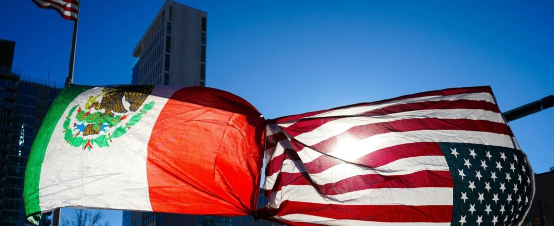A Mexican flag and a United States flag knotted together at their corners, flying above a city street against a clear blue sky with office buildings behind