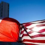 A Mexican flag and a United States flag knotted together at their corners, flying above a city street against a clear blue sky with office buildings behind