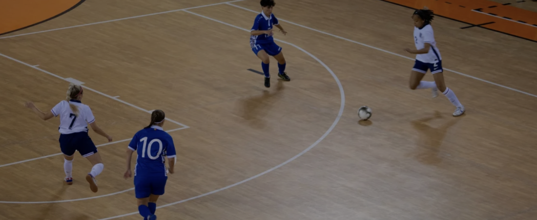 Players in blue and white kit chase a futsal ball on an indoor wooden court during a match