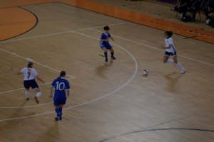 Players in blue and white kit chase a futsal ball on an indoor wooden court during a match