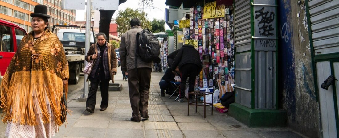 A woman in a bowler hat, fringed shawl and pleated skirt walks along a La Paz sidewalk past parked vehicles, other pedestrians and a stall selling phone cases