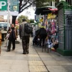 A woman in a bowler hat, fringed shawl and pleated skirt walks along a La Paz sidewalk past parked vehicles, other pedestrians and a stall selling phone cases