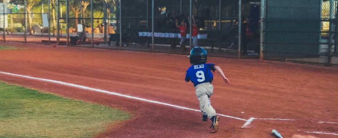 A young baseball player in a blue jersey numbered 9 runs along the first base line on a dirt infield, with teammates watching from a fenced dugout