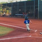 A young baseball player in a blue jersey numbered 9 runs along the first base line on a dirt infield, with teammates watching from a fenced dugout
