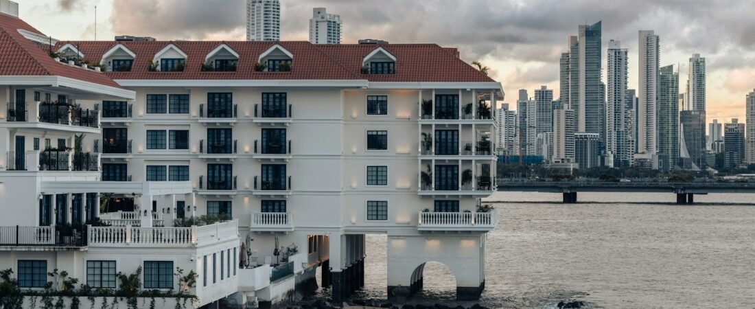 A white colonial building with a red tiled roof at the water's edge in Casco Viejo, Panama City, with the high rise towers of the modern financial district across the bay behind it