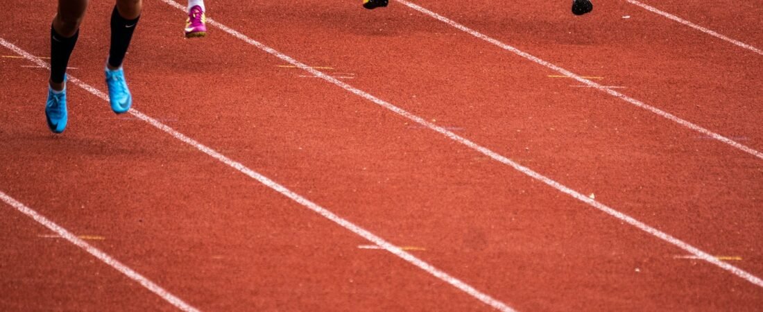 Three runners seen from the knees down, mid-stride in adjacent lanes on a red outdoor running track marked with white lines
