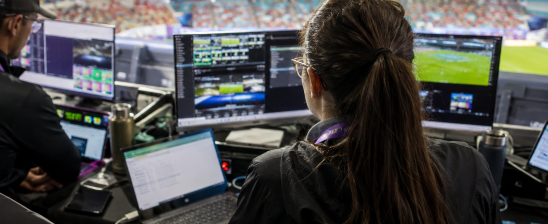 A production operator works at a bank of monitors showing camera feeds in a broadcast booth overlooking a stadium pitch