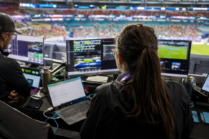 A production operator works at a bank of monitors showing camera feeds in a broadcast booth overlooking a stadium pitch