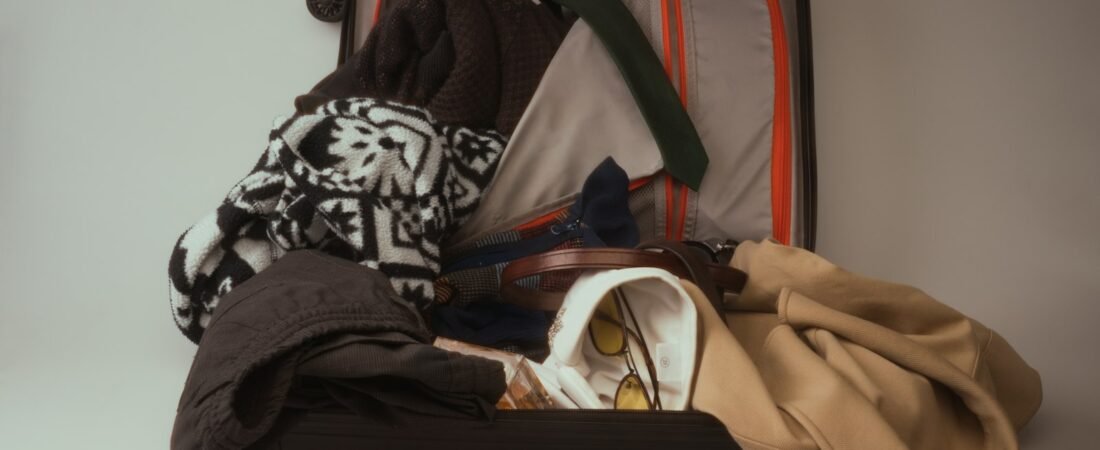 An open black hard-shell suitcase overfilled with a patterned sweater, dark jackets, a beige coat, a leather belt and a pair of sunglasses