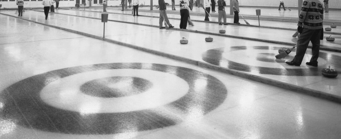 Concentric circles of a curling house marked under the ice, with stones resting on neighbouring sheets and a player standing with a broom