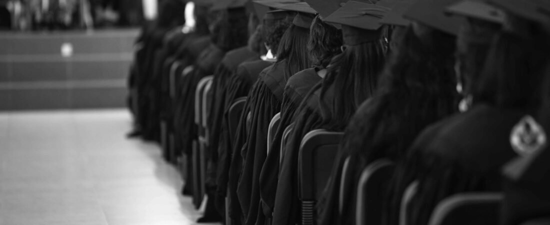 Rows of graduates in caps and gowns seated in a hall, photographed from behind