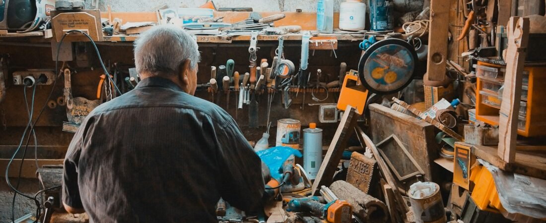 An older man seen from behind works at a cluttered wooden workbench, with hand tools hanging on the wall above and paint tins and clamps stacked around him