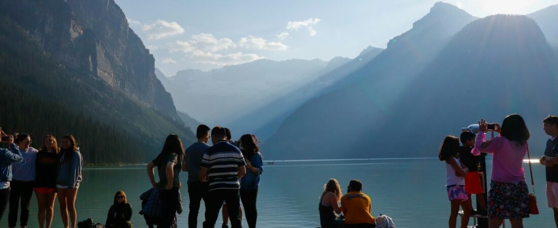 Around twenty visitors standing and sitting on a wooden lakeshore dock, backlit by low sun, with steep mountains rising on both sides of the lake behind them
