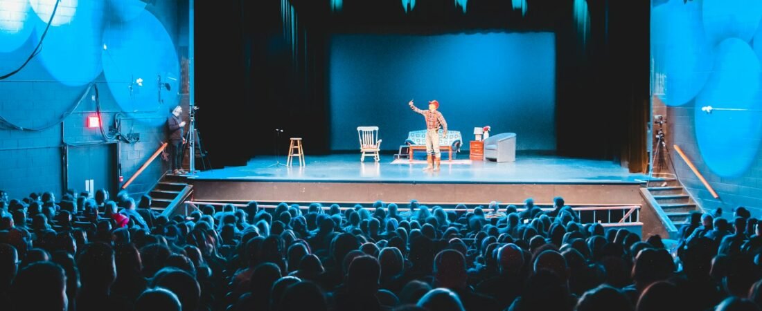 A full theatre audience seen from behind, facing a lit stage with a single performer, a white chair and a stool