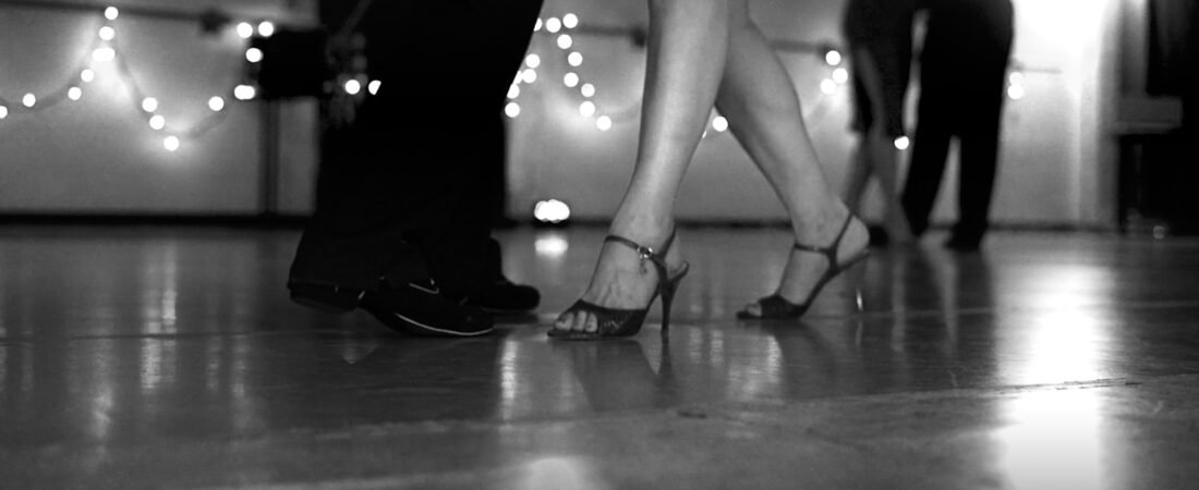 Low angle view of two dancers feet in dress shoes and heels on a dance floor, with string lights and other dancers blurred behind them