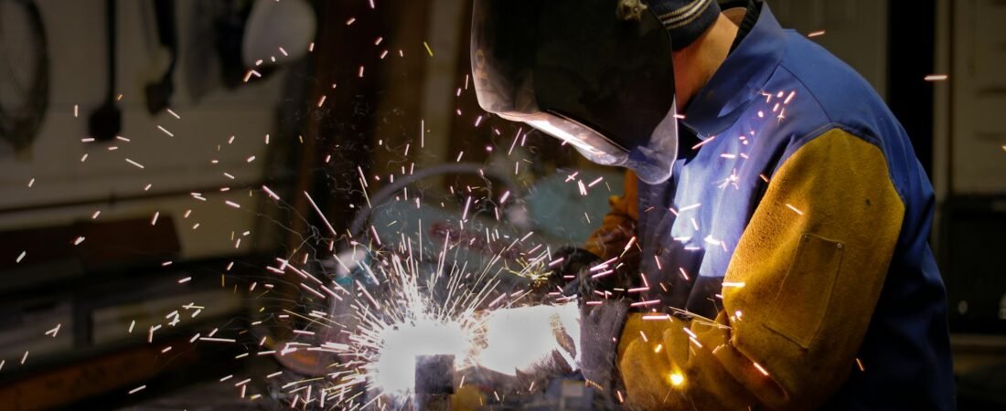 A welder in a dark full face hood and leather sleeves strikes an arc on a metal piece clamped to a bench, sparks scattering across a workshop wall hung with tools