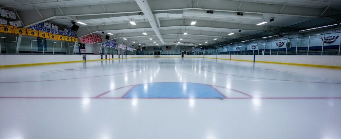 Wide view of an empty indoor ice rink with a blue centre-ice circle, white boards and banners along the sides